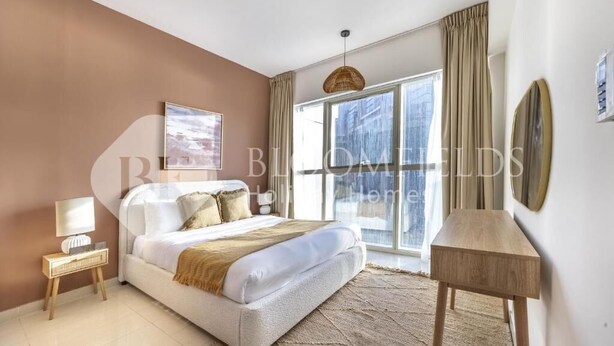 A bedroom with a large bed, a bedside table, a lamp, and a view of the city through large windows. - Bloomfields Iconic 1Br In Marina (Abu Dhabi)