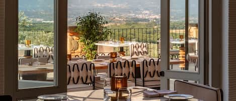 A dining area with a table and chairs, a view of a landscape through an arched window, and a large umbrella providing shade.