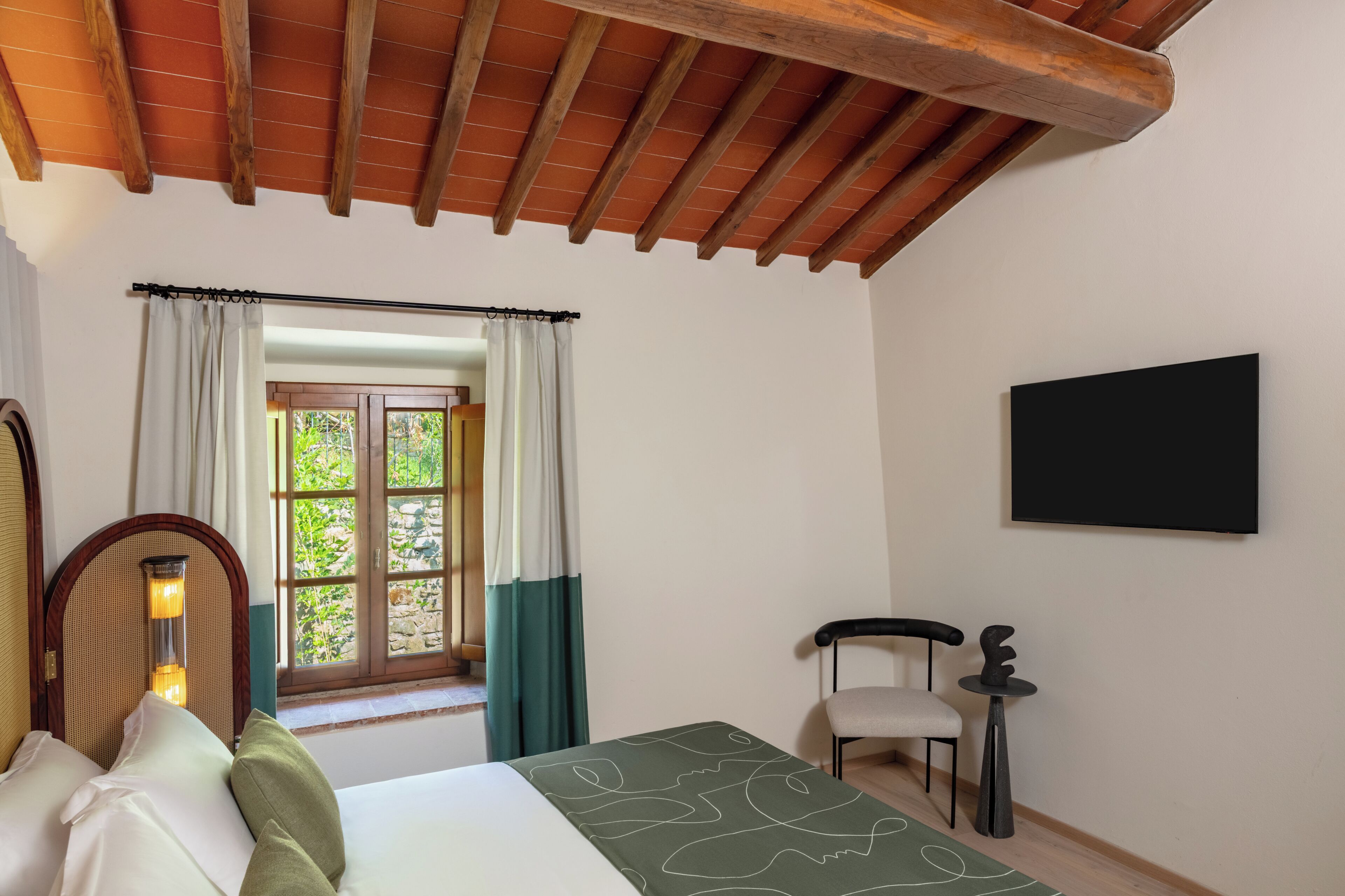 A room with a bed, a chair, a table, a window with curtains, and a flat-screen TV mounted on the wall.
