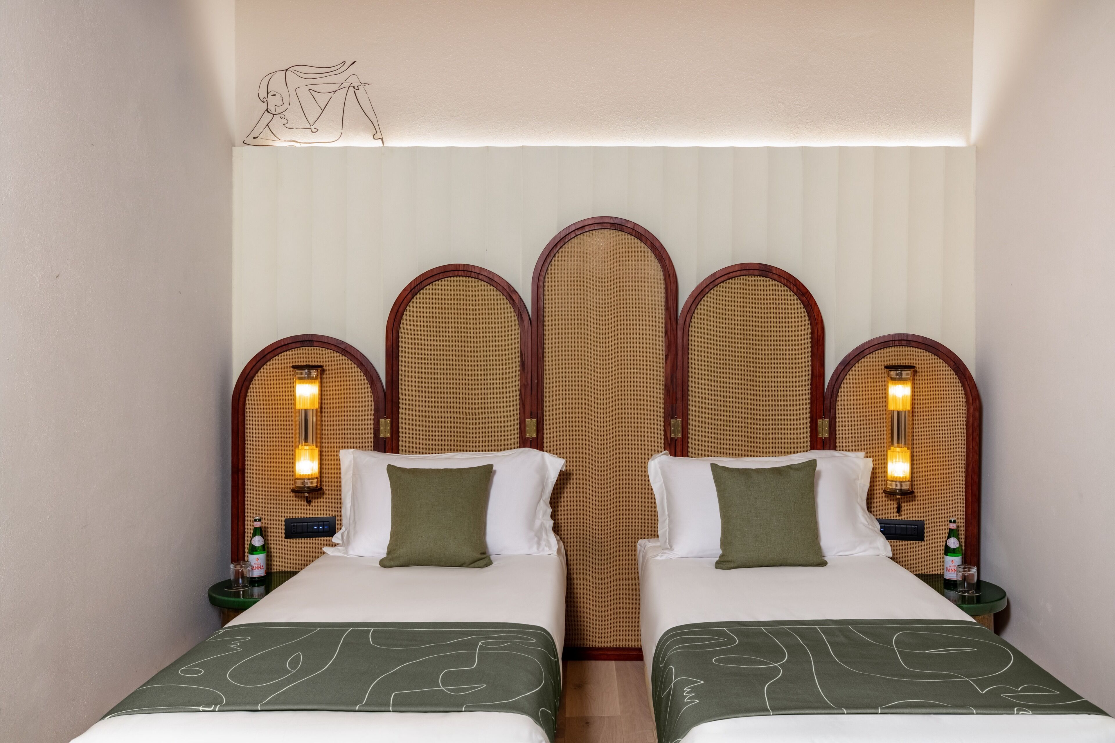 A hotel room with two beds, a decorative headboard, bedside lamps, and a bottle of water on the nightstand.