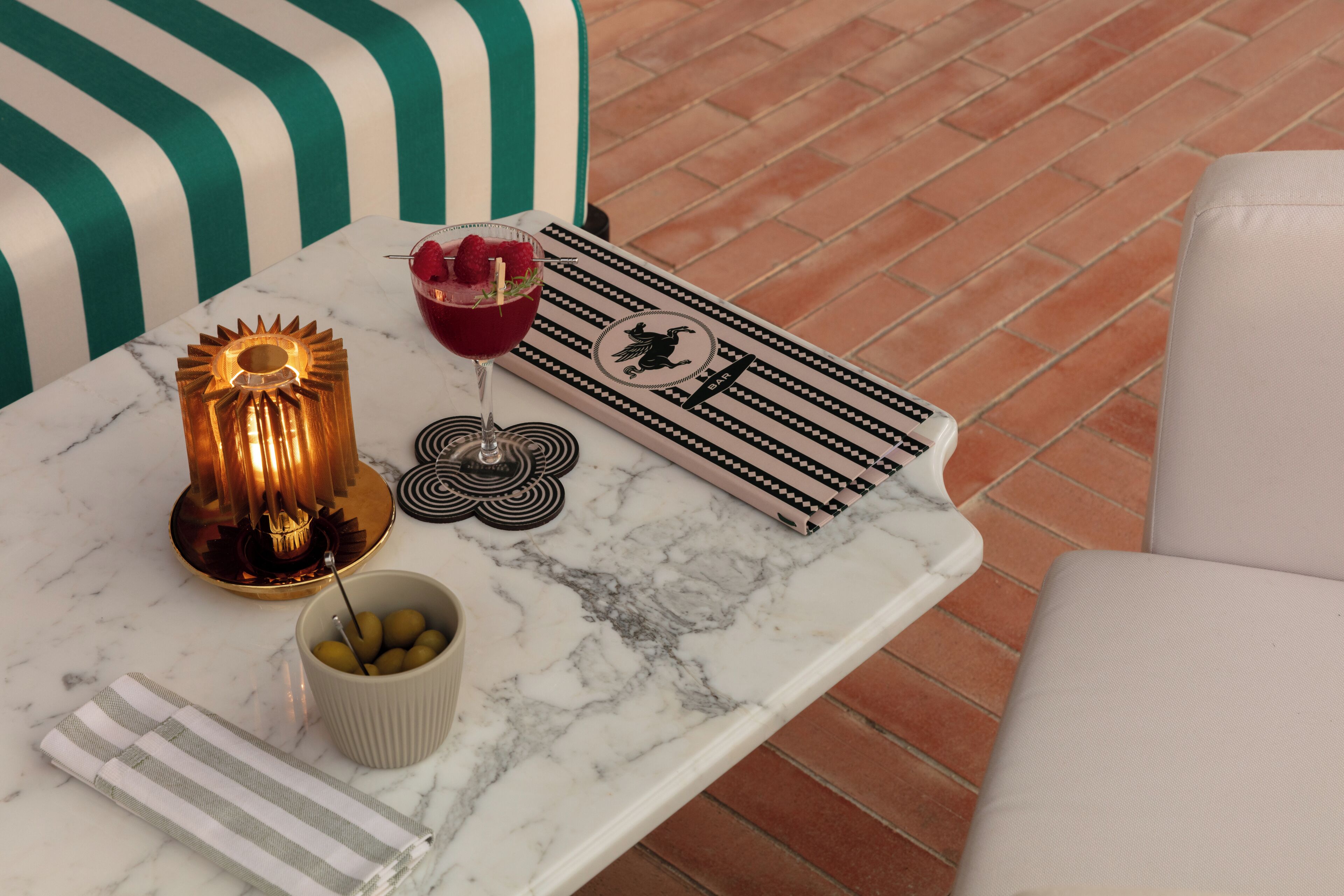 Uma mesa de mármore com uma lâmpada decorativa, um copo de bebida vermelha com framboesas, uma tigela de azeitonas e um guardanapo riscado.