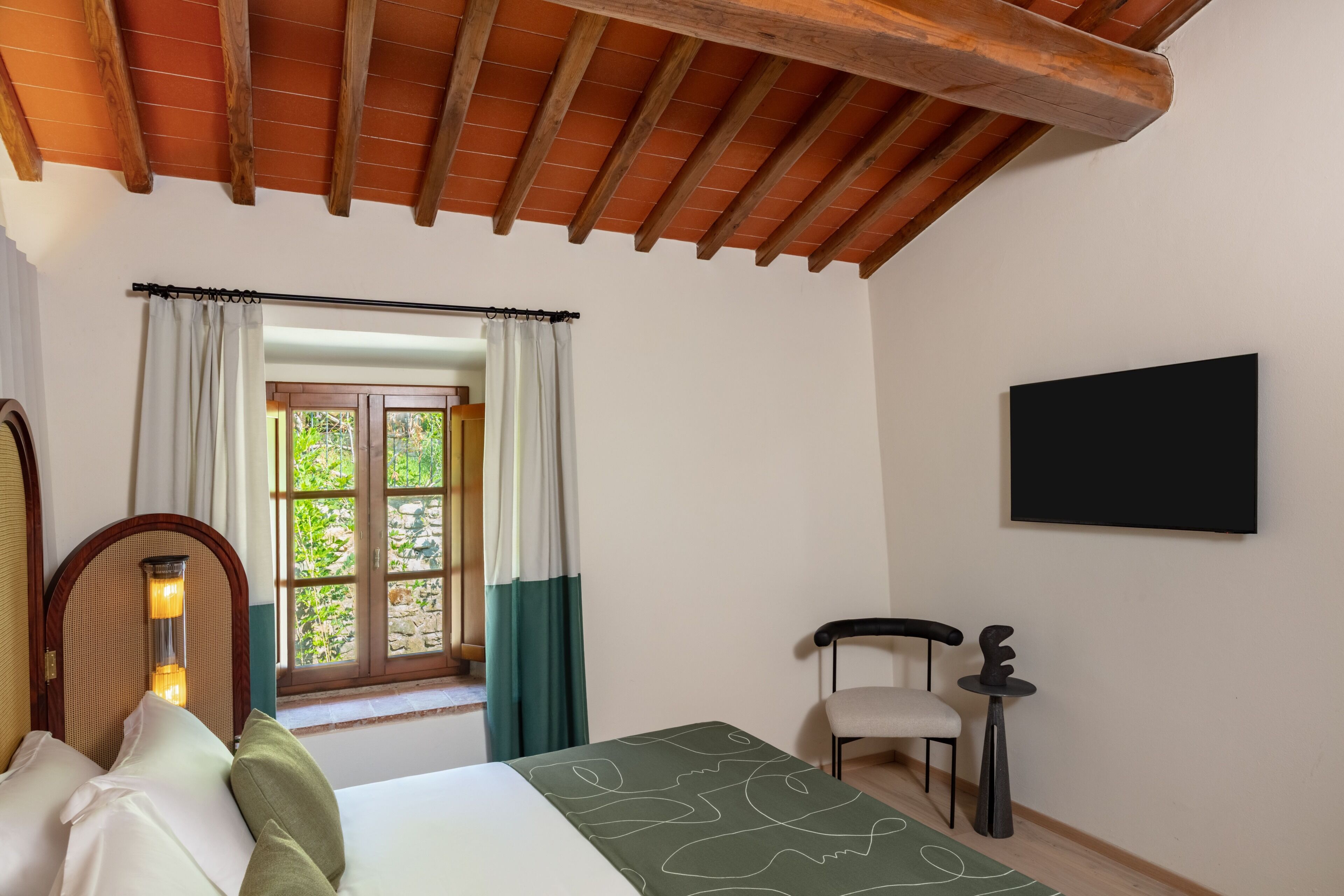 A bedroom with a bed, a chair, a table, a window with curtains, and a wall-mounted TV.