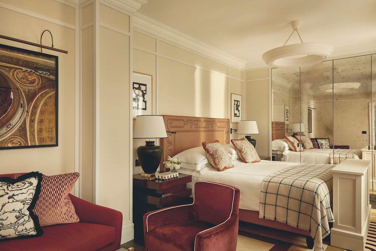 A hotel room with a large bed, a red armchair, a nightstand with a lamp, and a mirror.