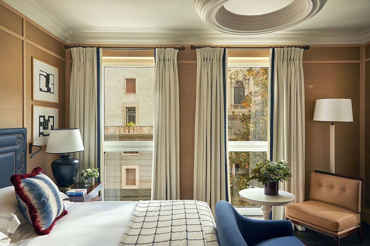 A hotel room with a bed, a chair, a table, a lamp, and a view of a building through large windows.