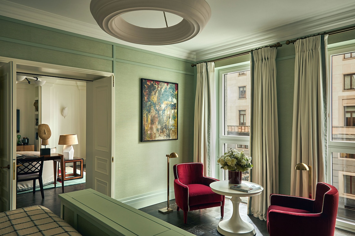 A hotel room with a large window, a red armchair, a white table, and a painting on the wall.