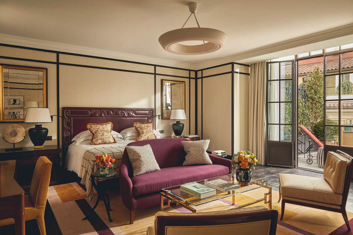 A hotel room with a large bed, a sofa, a coffee table with books, a chair, and a view of the outside.