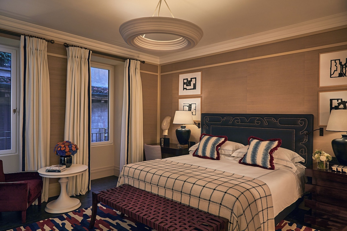 A hotel room with a large bed, a bedside table with a lamp, a chair, a window with curtains, and artwork on the walls.