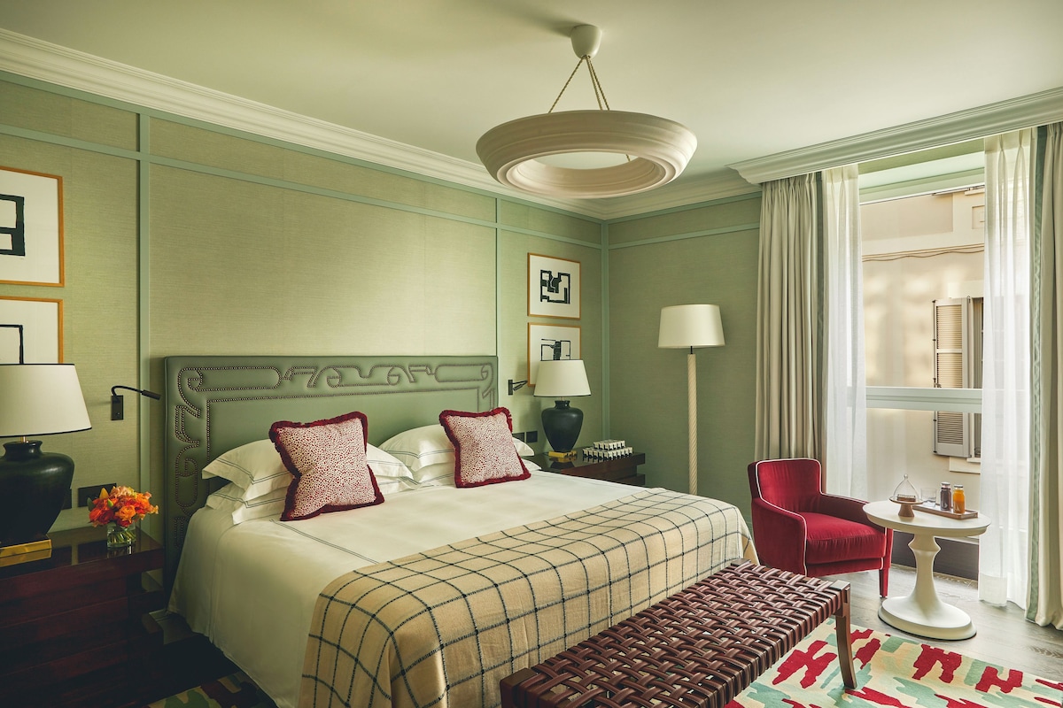 A hotel room with a large bed, a red armchair, a small table with a lamp, and a view of the outdoors through a window.