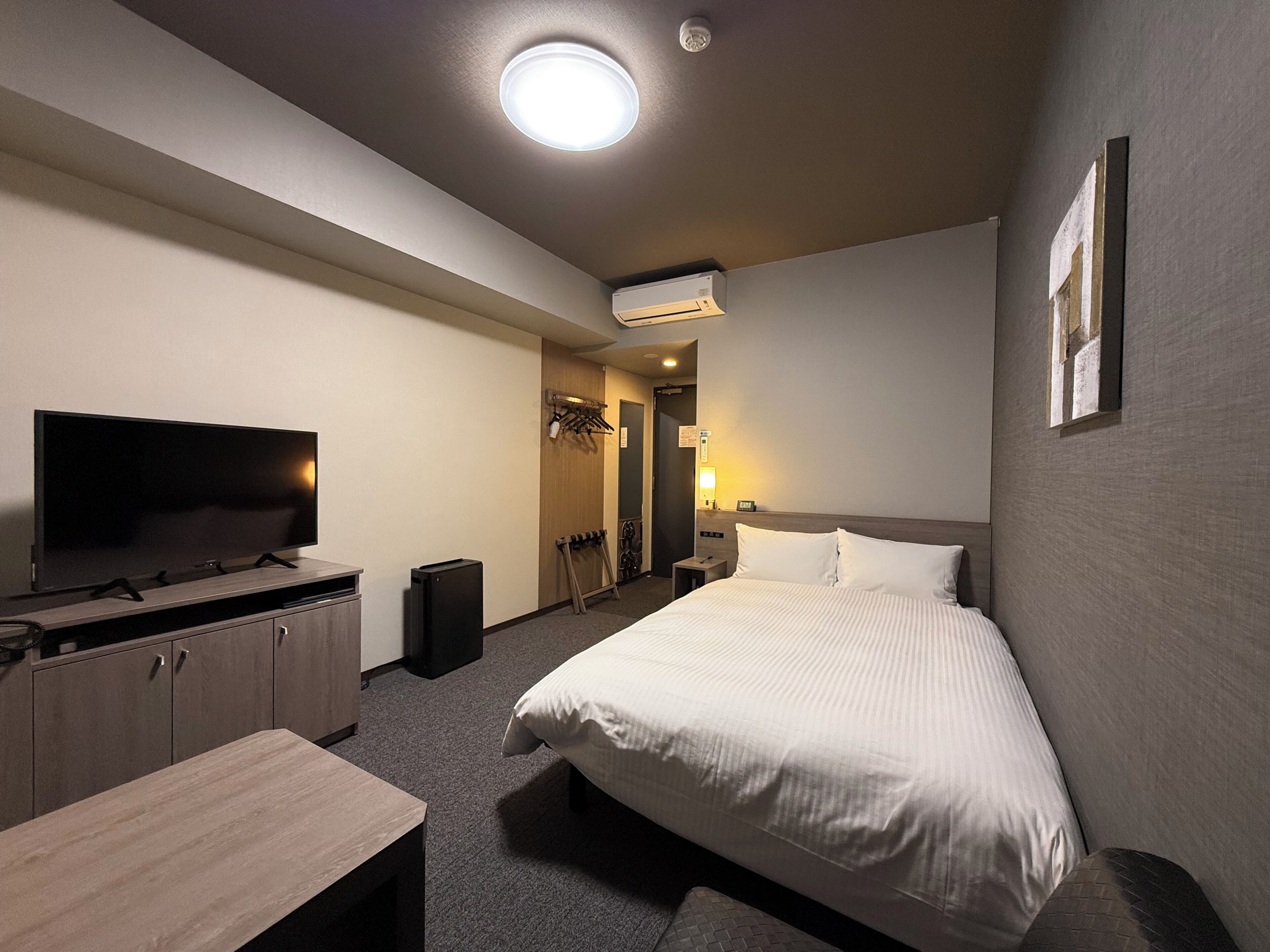 A hotel room with a bed, a TV, a desk, a wardrobe, and an air conditioning unit.