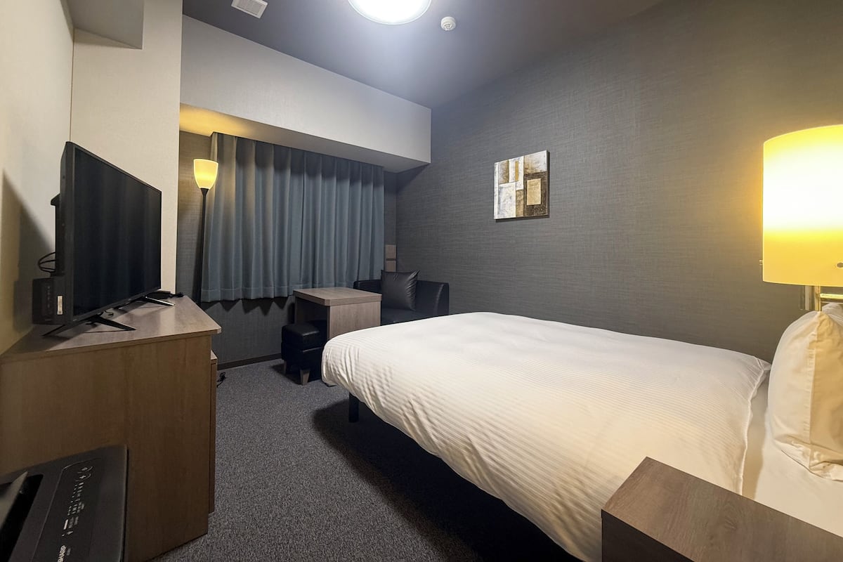 A hotel room with a bed, a desk with a computer, a chair, a lamp, and a ceiling light.