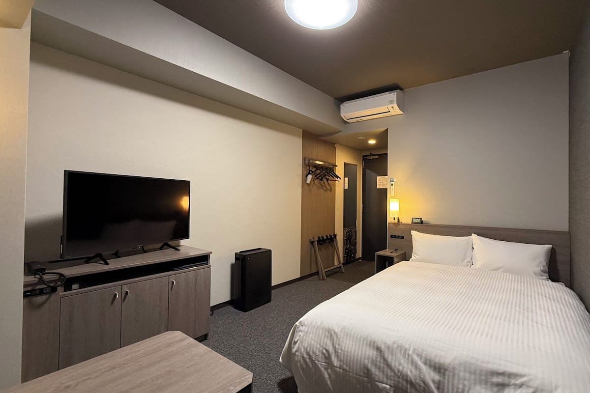 A hotel room with a bed, a TV, a desk, a wardrobe, a coat rack, and an air conditioning unit.