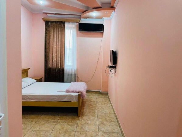 A small bedroom with a single bed, a window with curtains, an air conditioning unit, and a wall-mounted TV. - Aero Hotel (Parakar)