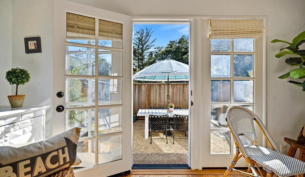 A room with a large glass door leading to an outdoor patio with a table and chairs, a potted plant, and a wicker chair.