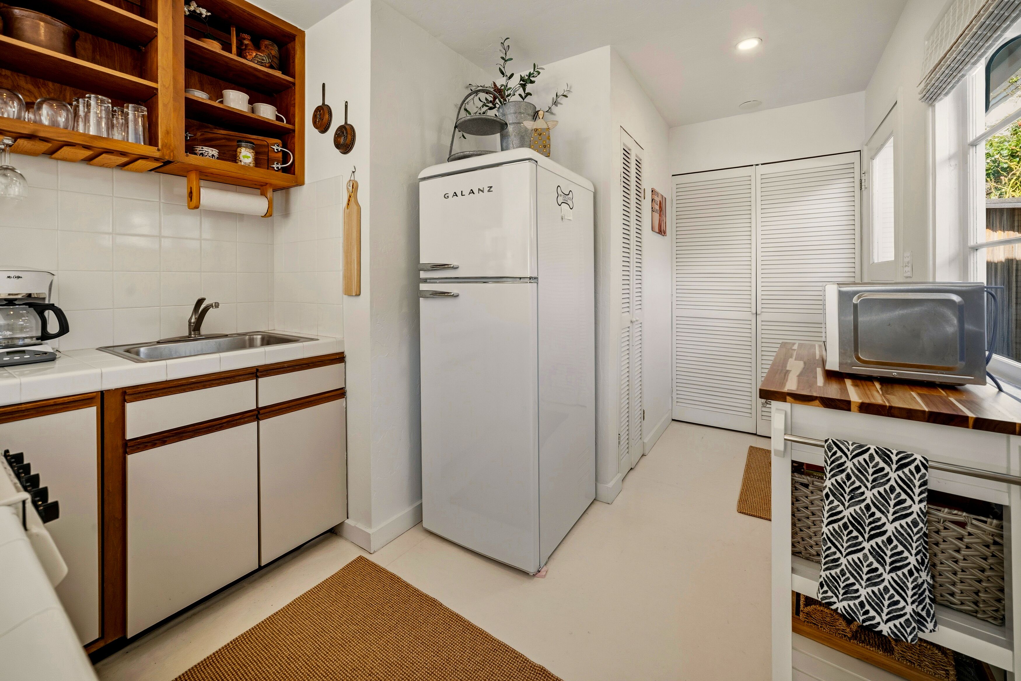 Una cocina compacta con armarios blancos, un lavamanos integrado, un refrigerador Galanz, una repisa de madera con utensilios de cocina y un televisor sobre un mueble de madera.