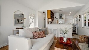 Una sala de estar moderna con un sofá blanco, una mesa de centro y una zona de cocina con armarios blancos y un estante de madera.
