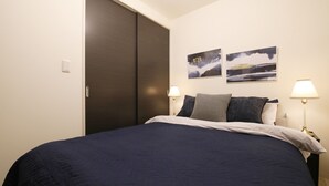Un dormitorio con una cama de color oscuro, dos lámparas fijadas a la pared y dos cuadros de paisajes.