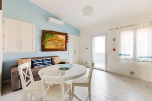 A dining area with a round table and white chairs, a sofa with patterned cushions, a painting on the wall, and a window with sheer curtains. - With private terrace, 50m from the sea (Castelsardo)