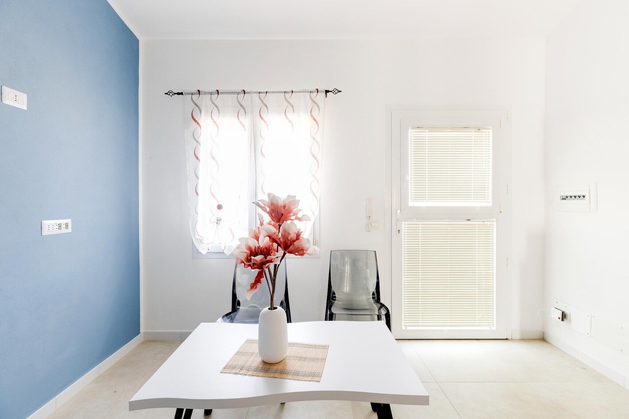 Ein Zimmer mit einem weißen Tisch, einer Blumenvase, einem Stuhl und einem Fenster mit Jalousien.