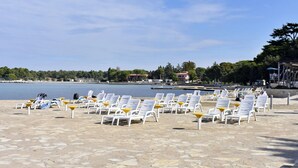 A beach with rows of white lounge chairs and yellow umbrellas, a calm sea, and a distant building. - Garden Apartment in Umag (Umag)