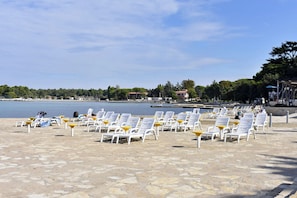Ein Strand mit Reihen aus weißen Liegestühlen und gelben Sonnenschirmen, einem ruhigen Meer und einem Gebäude in der Ferne.