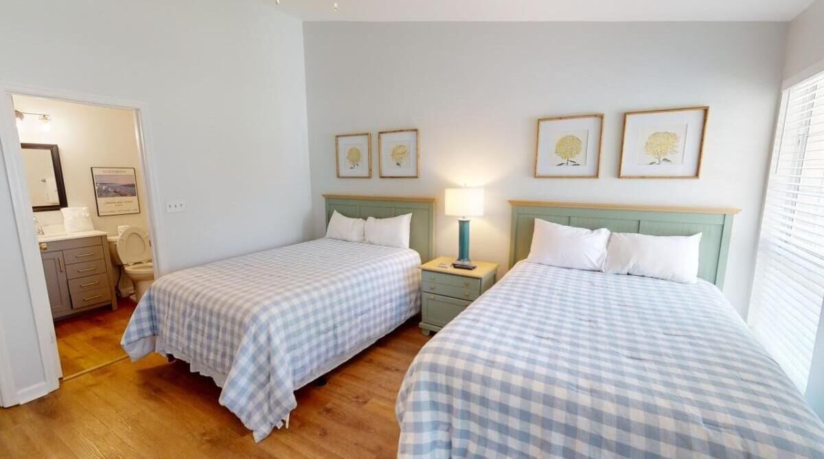 Une chambre d’hôtel avec deux lits, une petite table de chevet avec une lampe, une salle de bain visible à travers une porte ouverte et des tableaux encadrés accrochés au mur.