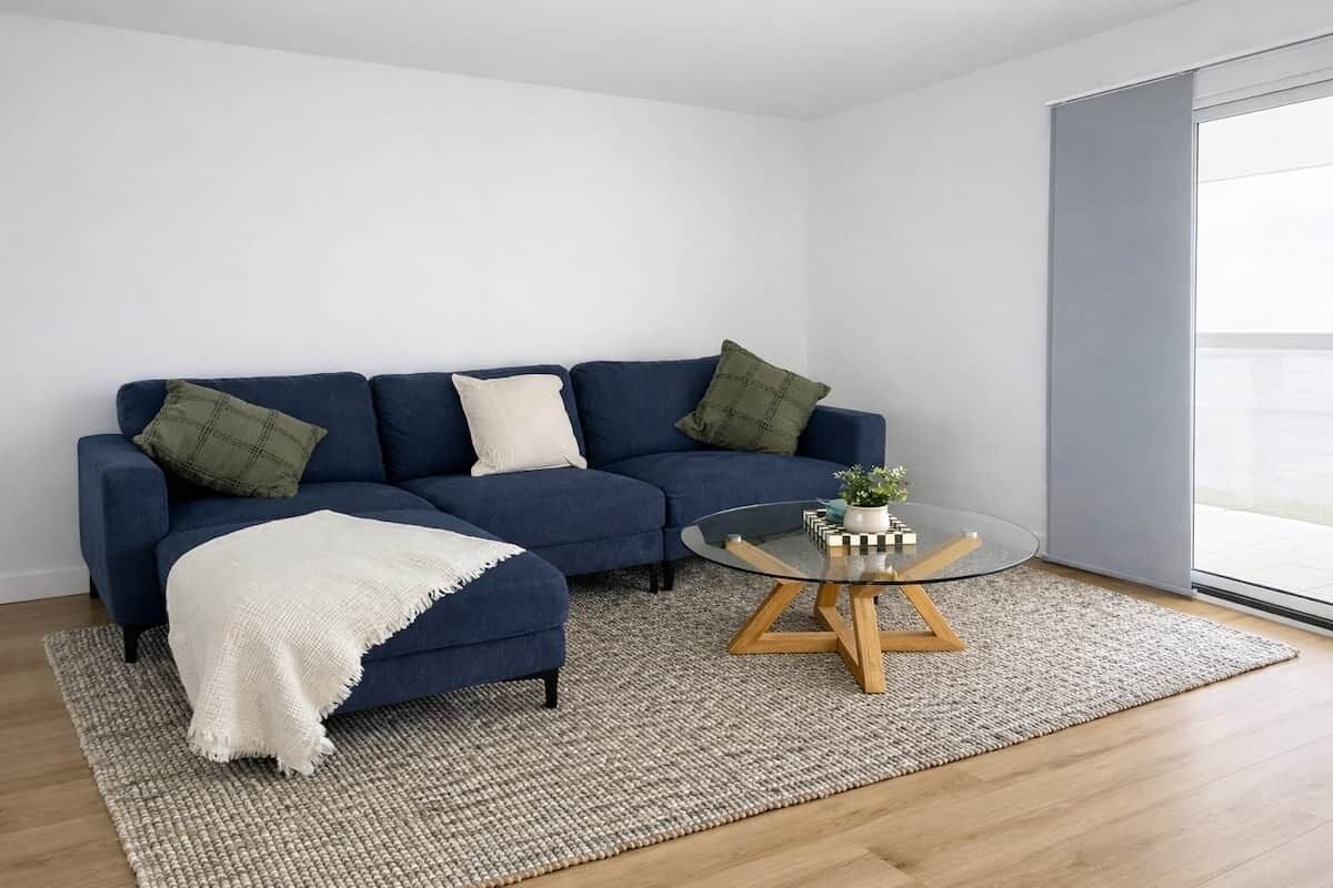 Una sala moderna con un sofá azul, una mesa de centro de vidrio y un ventanal grande.