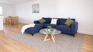 A modern living room with a blue sofa, a glass coffee table, and a dining area with wooden chairs. - 'Bentwood Escape' 
Modern unit in heart of Yamba (Yamba)