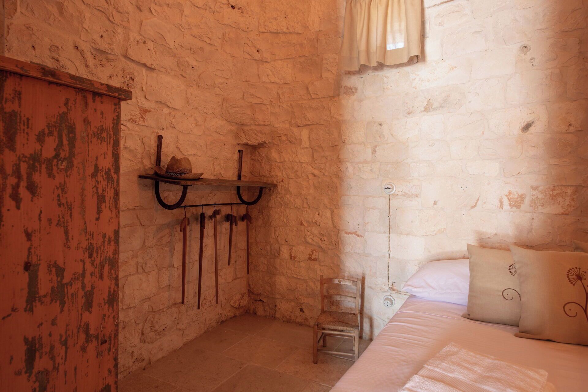 Una habitación rústica con una cama, una silla de madera, una estantería con un sombrero y un gancho en la pared.