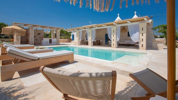 Area piscina con poltrone a sdraio, muro in pietra e zona relax coperta con tende bianche.