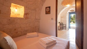 Una stanza con una parete in pietra, un letto con asciugamani piegati, un quadro appeso e vista su uno spazio esterno con piscina e sedie.