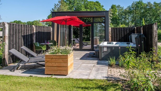 Un patio trasero con terraza de madera, jacuzzi, sombrilla roja y cerramiento de vidrio.