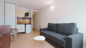 En kompakt opholdsstue med et tekøkken, en grå sofa, et lille hvidt bord og en trækommode.