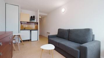 Ein gemütlicher, kompakter Wohnraum mit einer Küchenzeile, einem grauen Sofa, einem kleinen weißen Tisch und einer Holzkombination.
