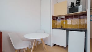 Et kompakt tekøkken med mikrobølgeovn, kaffemaskine og køleskab. Der er et lille hvidt bord med to stole.