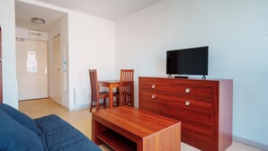A compact living space with a wooden TV stand, a flat-screen TV, a dining table with chairs, and a blue sofa. - Lush corner near the local bistro (Agde)