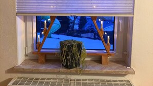 Een raam met uitzicht op een besneeuwd landschap, een potplant en kaarsen op een houten standaard.