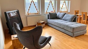 A modern living room with a grey sofa, a grey armchair, and a wooden coffee table. There are three windows with wooden frames and a view of a building. A television is on a stand to the left. - Haus De Kleene & Gruusse in Mildenau (Mildenau)