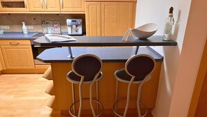 A kitchen with wooden cabinets, a black countertop, a coffee machine, and two bar stools. - Haus De Kleene & Gruusse in Mildenau (Mildenau)