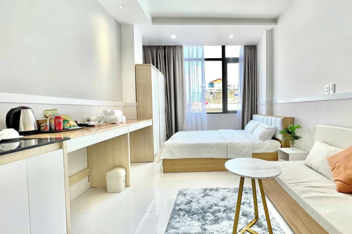 A modern hotel room with a bed, a small table, a sofa, a kitchen area, and a view of the outside.