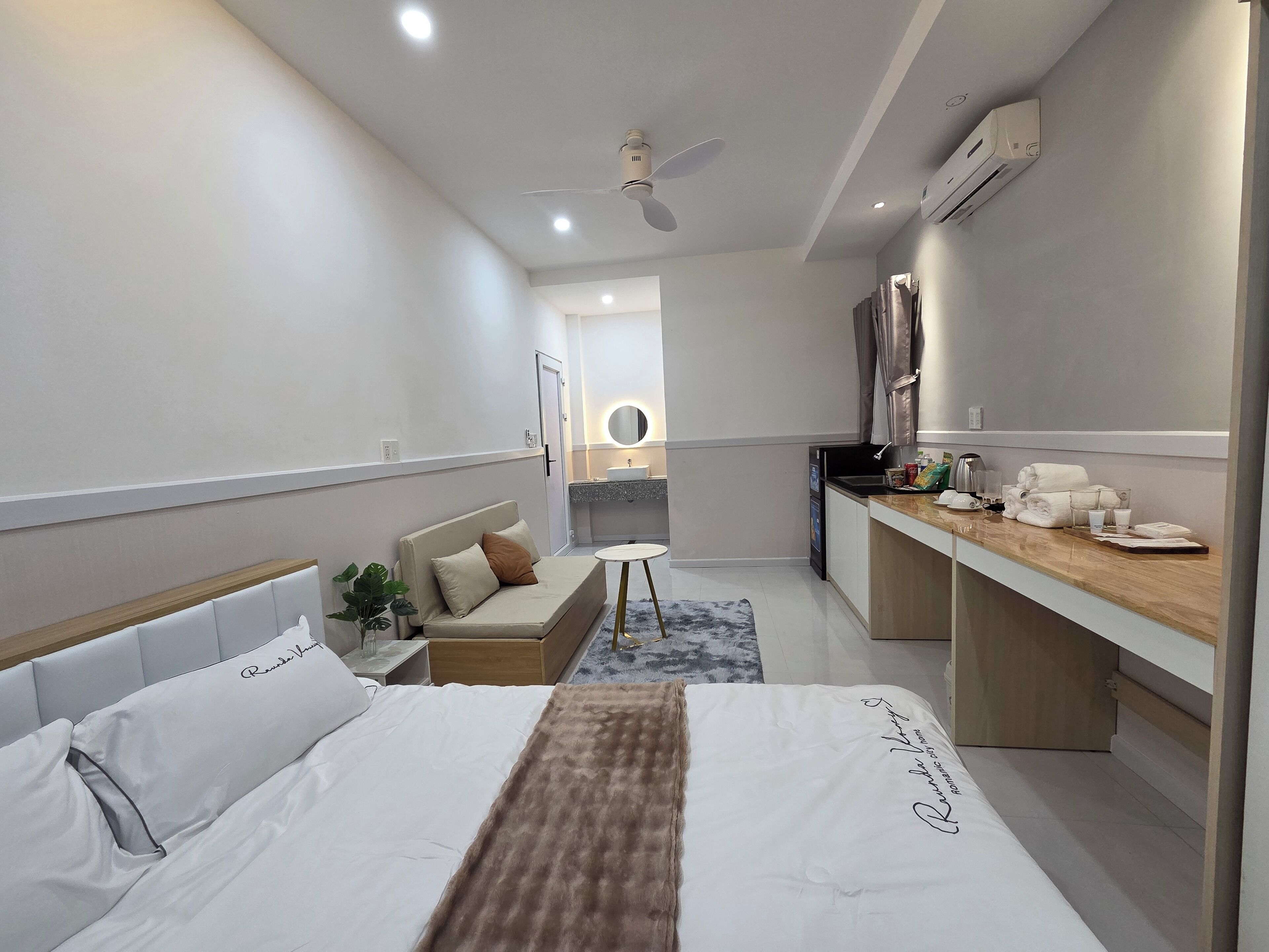 A modern hotel room with a bed, a sofa, a small table, a kitchenette, and a ceiling fan.