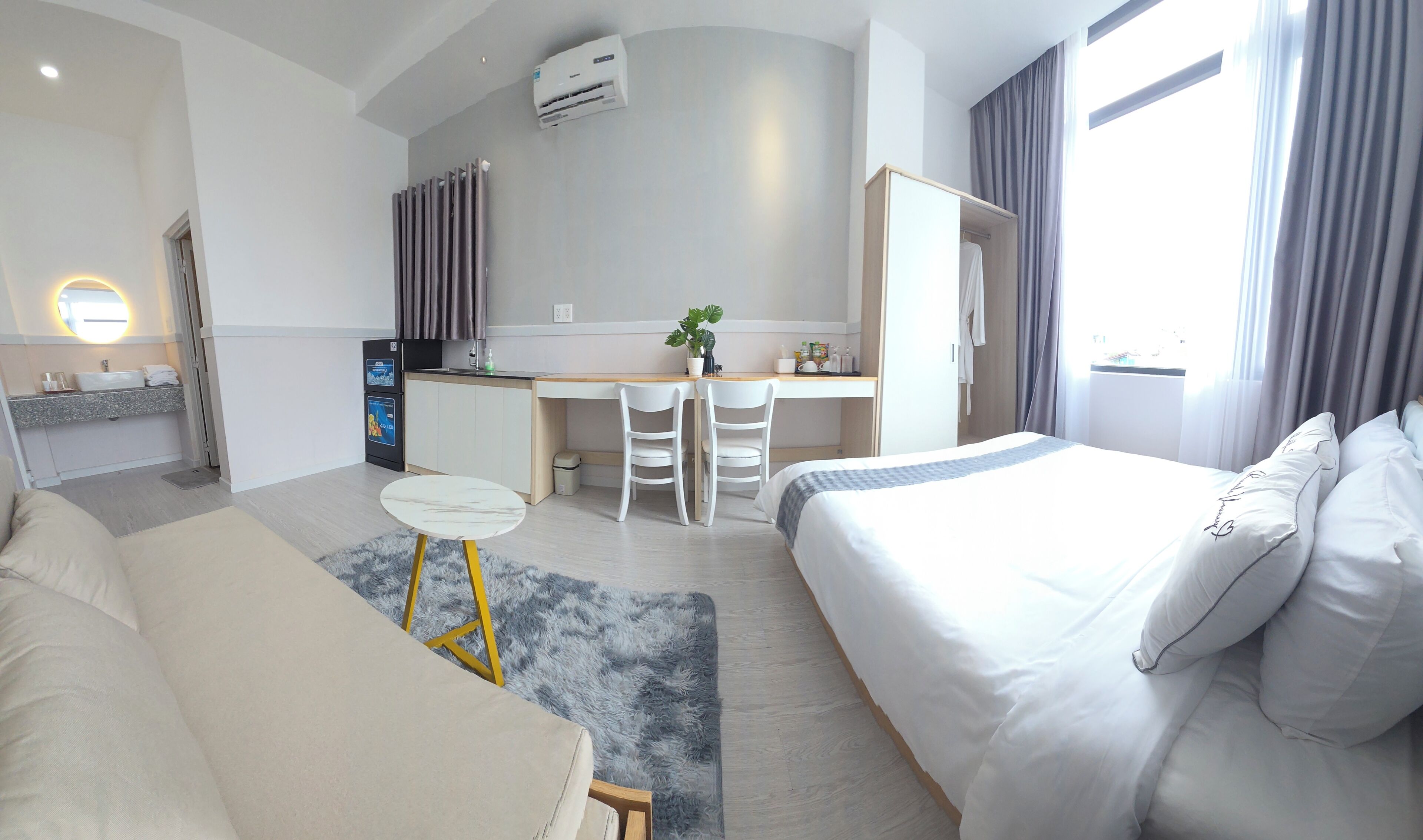 A hotel room with a bed, a desk, two chairs, a small table, a sofa, a bathroom with a sink, and a window with curtains.