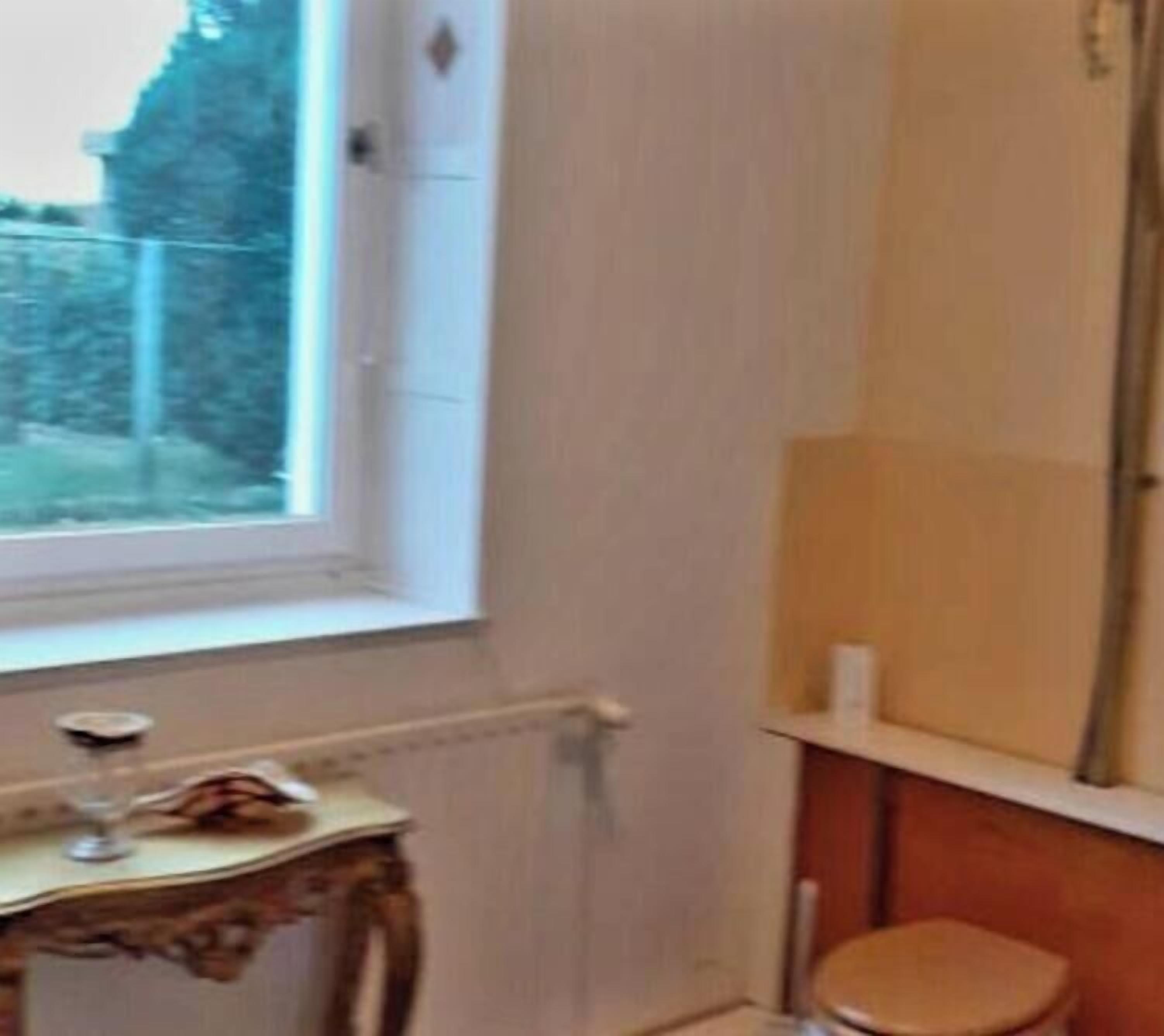 Ein Zimmer mit Fenster, Heizkörper, einem kleinen Tisch mit einem Glas und einem hölzernen Hocker.