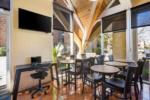 Un área de estar interior con mesas y sillas, un televisor de pantalla plana montado en la pared y una pared de ladrillo.