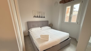 Ein ordentlich bezogenes Bett mit grauem Kopfteil, eine weiße Wand mit einem gerahmten Bild und ein Fenster mit Blick auf Gebäude.