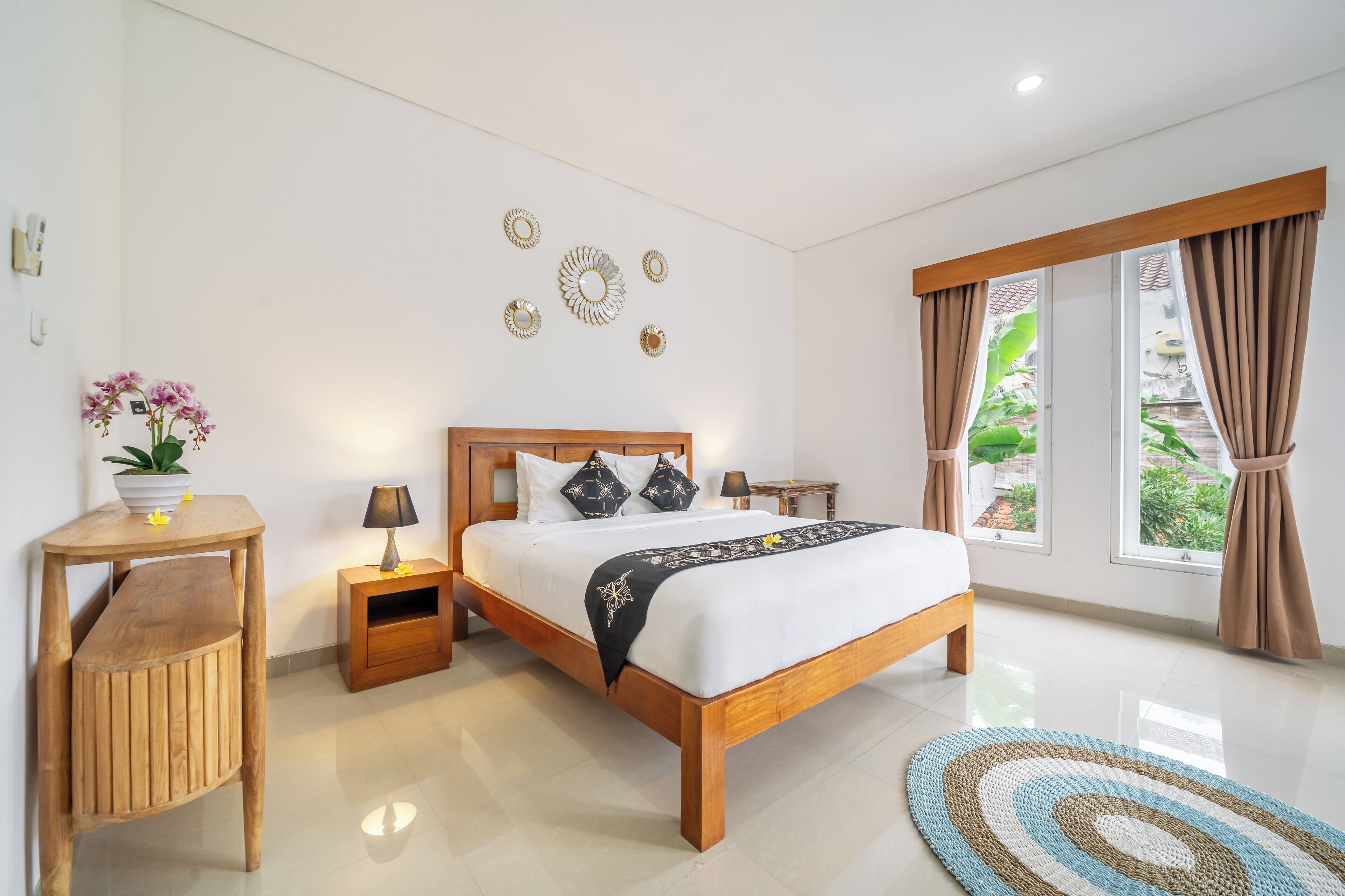 A bedroom with a wooden bed, bedside tables, a nightstand, a wall-mounted lamp, decorative wall art, and large windows with curtains.