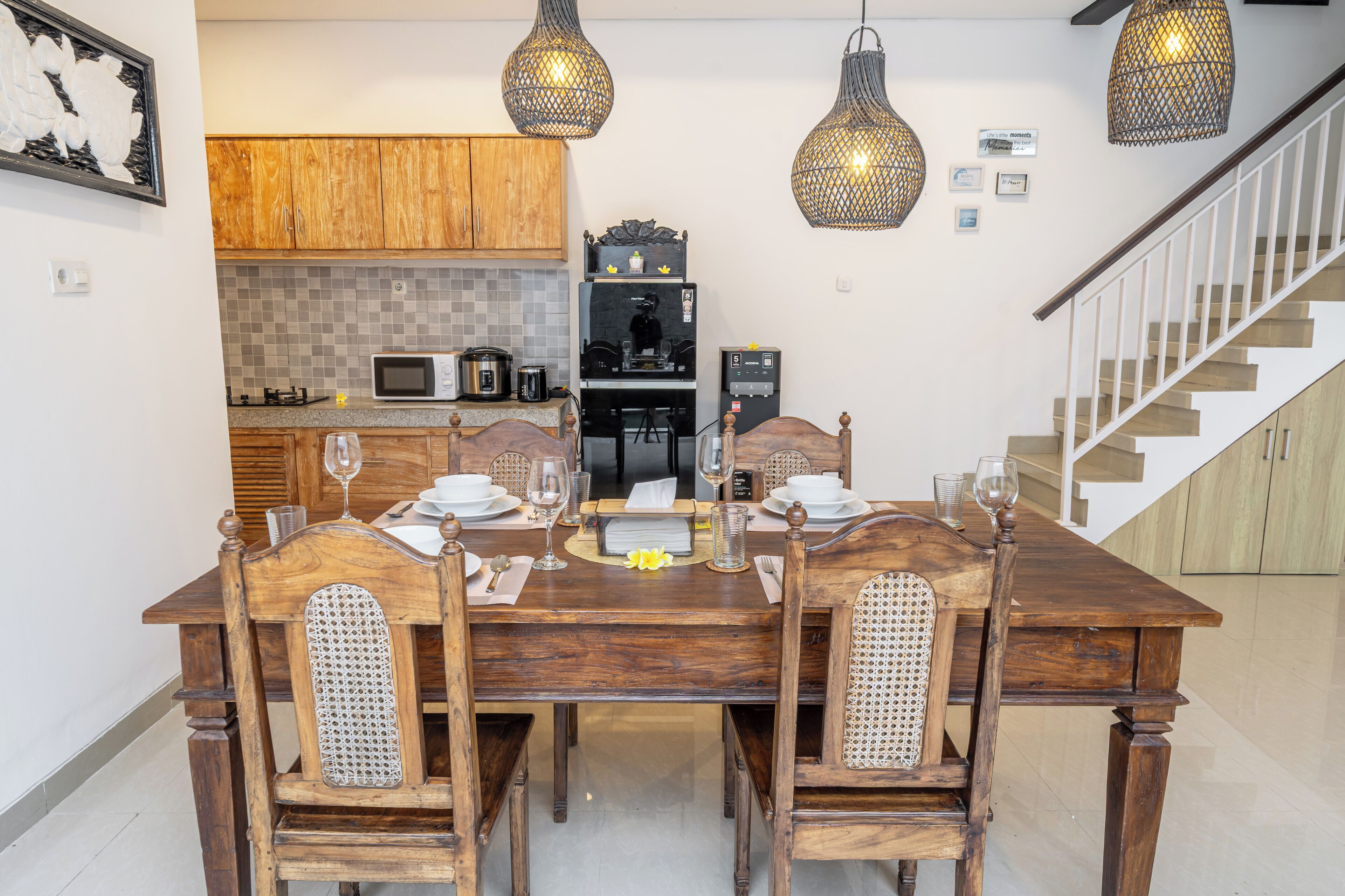 A dining area with a wooden table set for four, surrounded by wooden chairs with woven backrests. A kitchen with wooden cabinets and a microwave is visible in the background.