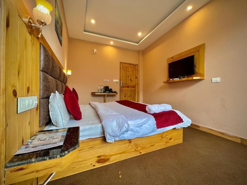 Un dormitorio con cabecera de madera, cama con ropa de cama blanca y roja, una mesita de noche pequeña con una lámpara, y un televisor montado en la pared.