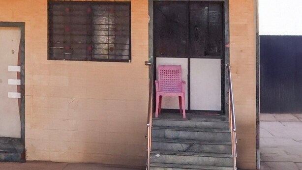 A building with a beige exterior, a set of stairs leading to a door, and a pink chair in front of the door. - Collection O Mahabaleshwar (Jaoli)