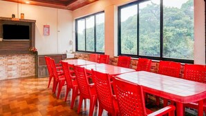 A conference room with red chairs and a table, large windows with a view of trees, a flat-screen TV on the wall, and a ceiling fan. - Collection O Mahabaleshwar (Jaoli)