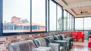 A modern lounge area with leather sofas, a brick accent wall, large windows, and a view of buildings outside. - Collection O Mahabaleshwar (Jaoli)
